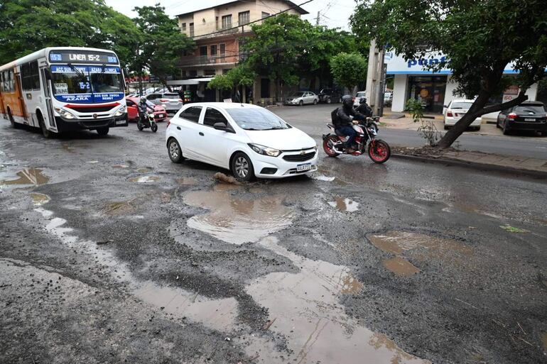 Automóvil blanco y autobús en avenida deteriorada, con baches y árboles de fondo; clima nublado en Vista Alegre.