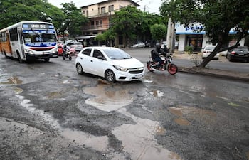 Automóvil blanco y autobús en avenida deteriorada, con baches y árboles de fondo; clima nublado en Vista Alegre.