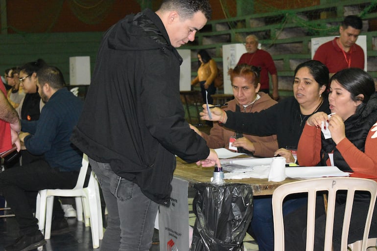 Hombre de pie con chaqueta negra interactuando con cuatro mujeres tras una mesa en un ambiente electoral.