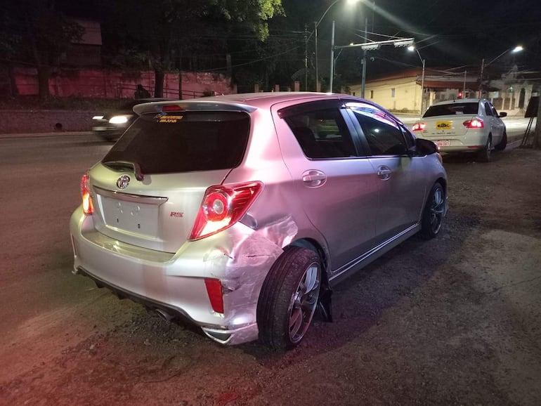 Automóvil hatchback plateado con daños visibles estacionado, luces traseras encendidas, mientras un taxi blanco está a su lado en un entorno urbano.