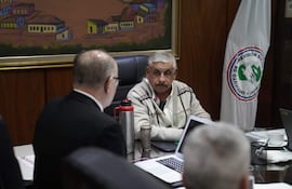 Isaías Ricardo Fretes, hombre mayor con suéter claro, dialoga con un hombre en traje con anteojos en una oficina colorida.
