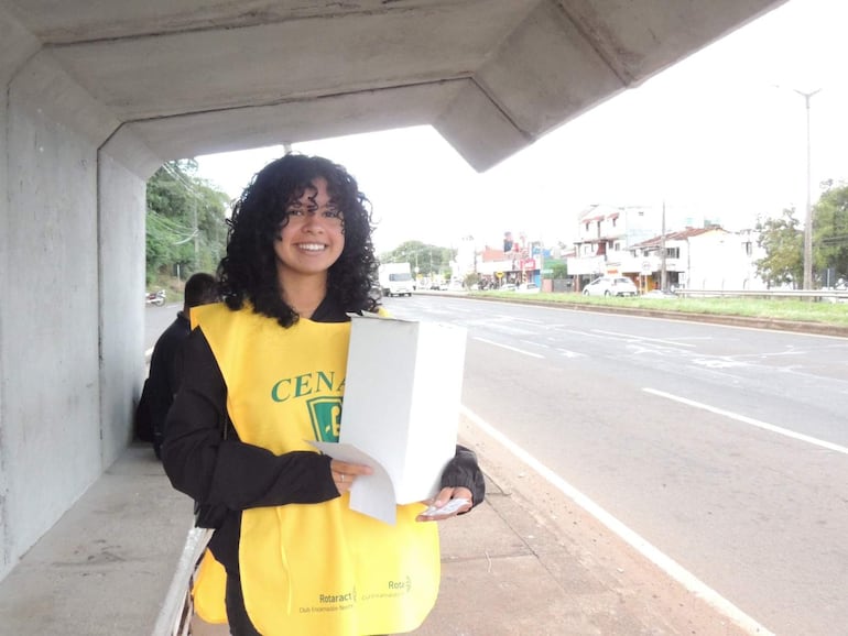 Joven con cabello rizado y chaleco amarillo sonríe, sosteniendo una caja blanca en una calle con edificios al fondo.