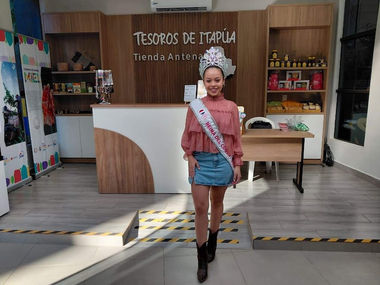 Mujer con corona y banda 'Reina del Cora' en vestido rosa y falda de mezclilla, en la tienda Tesoros de Itapúa.