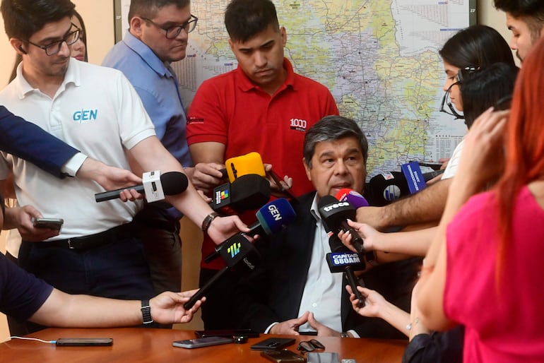 Hugo Arce en traje oscuro, sentado en mesa mientras responde preguntas de reporteros y cámaras en sala de prensa.