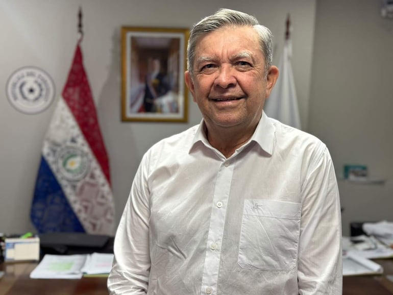 Arsenio Zárate, con cabello blanco y camisa blanca, sonríe en una oficina con banderas de Paraguay a su lado.