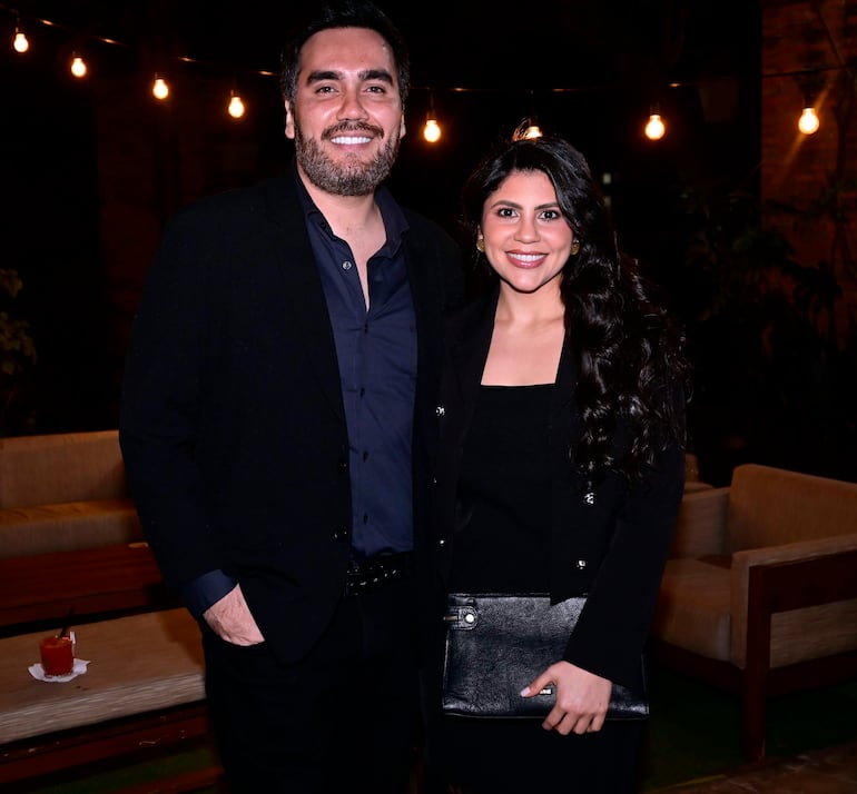 Hombre con camisa oscura y abrigo negro sonríe junto a mujer de blusa negra y abrigo oscuro, en ambiente acogedor con luces colgantes.