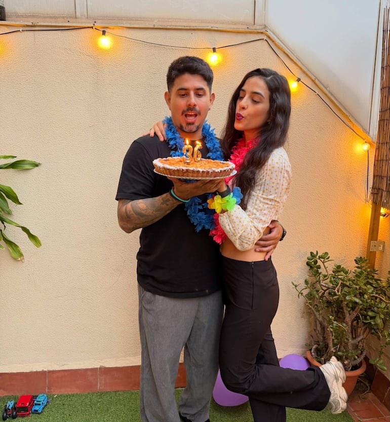 Hombre con camiseta negra y collar hawaiano sostiene un pastel, mientras una mujer sonríe con blusa blanca y guirnalda de flores.