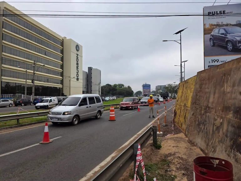 Vía urbana con furgoneta plateada en movimiento y auto rojo detenido, conos de tráfico y edificio Bourbon al fondo.