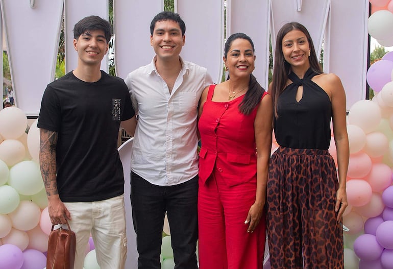Cuatro personas sonrientes, dos hombres con camisetas claras y dos mujeres en conjunto rojo y blusa negra, frente a decoraciones de globos pastel.