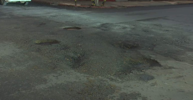 Calle en mal estado con baches visibles y un vehículo estacionado. Ambiente nocturno y falta de mantenimiento.