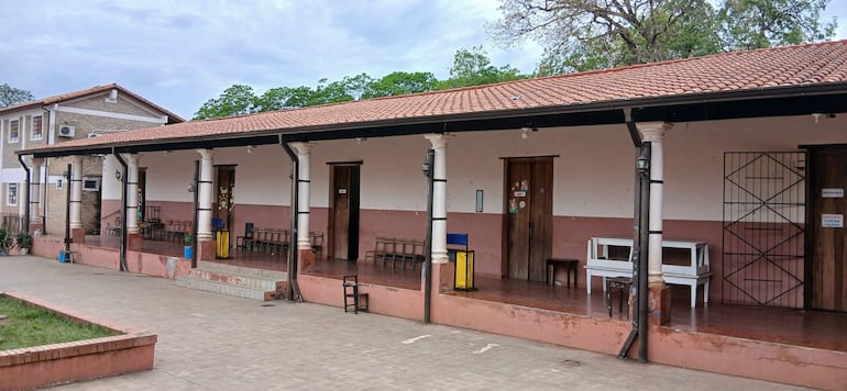 Edificio de una planta con techo deteriorado y carteles que indican 'BIBLIOTECA', rodeado de sillas de madera y árboles.
