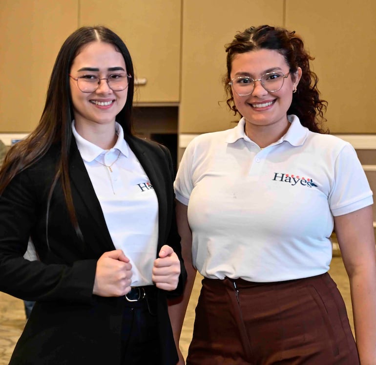 Mujeres sonrientes, una en camisa blanca y abrigo negro, otra en camiseta blanca con logo 'Hayes', en ambiente profesional.