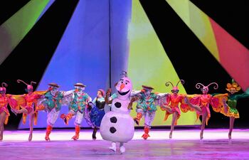 Personajes de Disney bailan en el escenario; uno en forma de muñeco de nieve, vestidos coloridos y alegres, ambiente festivo y luminoso.