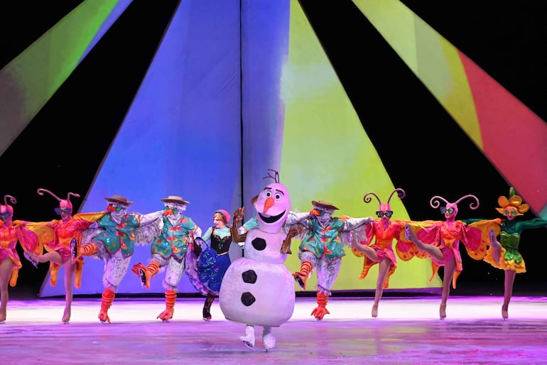Personajes de Disney bailan en el escenario; uno en forma de muñeco de nieve, vestidos coloridos y alegres, ambiente festivo y luminoso.