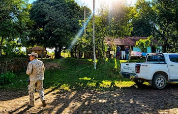 Hombre en uniforme camuflado camina por un camino de piedra, rodeado de vegetación y casas en un ambiente rural.