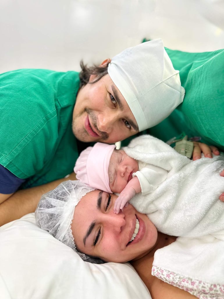 Padre en camiseta verde y cofia blanca sonríe, mientras madre sonríe sosteniendo a su bebé envuelto en manta blanca y gorro rosa.