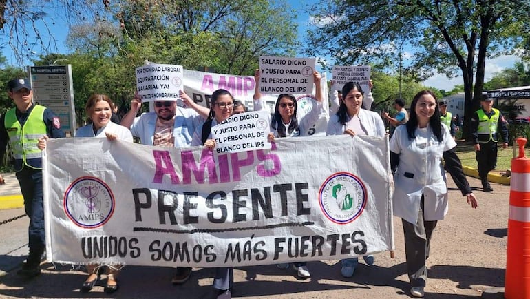 Grupo de médicos en camisas blancas sostiene carteles, entre ellos una mujer con el lema 'AMIPS PRESENTE', durante una protesta pacífica.
