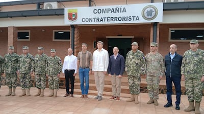 Grupo de ocho personas, cinco con uniformes militares y tres en ropa civil, posan frente a un edificio con paredes de ladrillo.