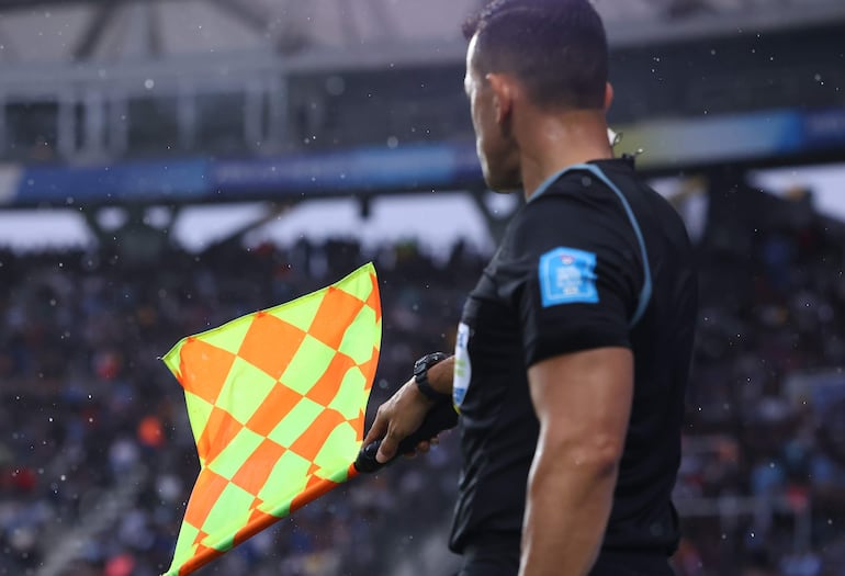 Árbitro con camiseta oscura y detalles azules, sosteniendo bandera amarilla y verde en ambiente lluvioso, rodeado de multitud.