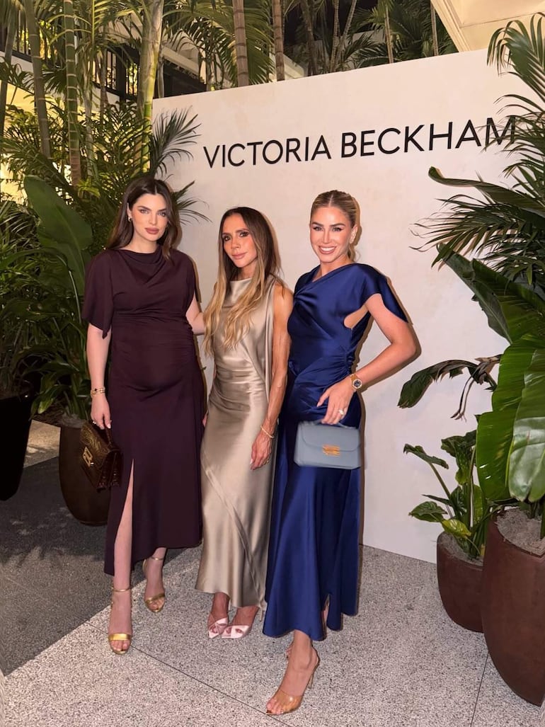 La imagen muestra a tres mujeres posando delante de un fondo que dice "VICTORIA BECKHAM". 

1. **Personas presentes**: Hay tres mujeres. Una está vestida con un vestido oscuro, otra con un vestido plateado y la tercera con un vestido azul. La mujer en el centro parece ser el foco, mientras que las otras dos están a su lado, en posiciones simétricas.
  
2. **Rol visual**: La mujer de vestido plateado está en el centro, interacciones relajadas con las otras dos, que están más a los lados. 

3. **Ambiente**: Se trata de un entorno exterior, posiblemente en un evento social, con plantas que rodean el área, sugiriendo un ambiente elegante y sofisticado.

No hay elementos relacionados con el fútbol ni otros contextos noticiosos visibles. No se identifican figuras públicas con alta confianza.