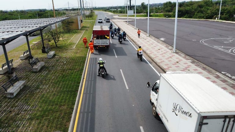 Trabajador en chaleco naranja supervisa obras viales sobre carretera con camiones y motos en movimiento.