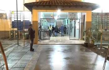 Hombre de pie con ropa oscura vigilando la entrada del colegio, mientras varias personas con uniforme se comunican dentro del edificio.