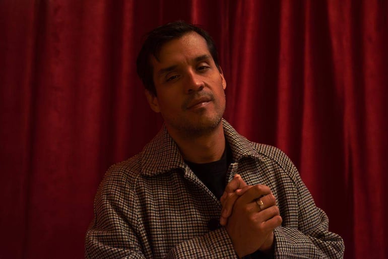 El David Aguilar lleva abrigo a cuadros, de pie con expresión tranquila, en un entorno con cortina roja de fondo.