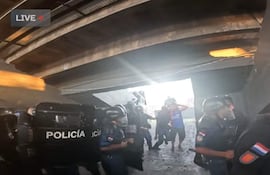 Agentes de policía con escudos en primer plano, controlando una situación tensa en un lugar cerrado con luz tenue.