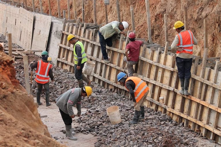 Seis trabajadores activos en la construcción, usando chalecos y cascos de colores, en un entorno de tierra y piedras.