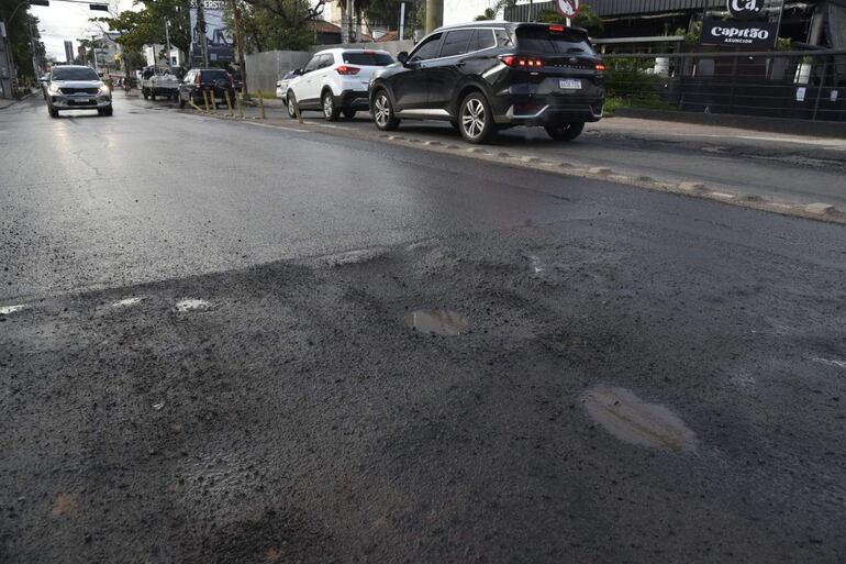 Calle de Asunción con bache en primer plano y vehículos en movimiento. Ambiente urbano deteriorado.