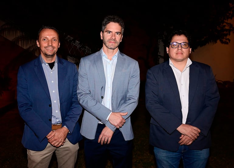 Tres hombres en chaquetas formales, con expresiones serias, posan al aire libre en ambiente tenue.