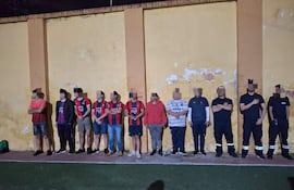 Diez personas alineadas contra una pared amarilla, vistiendo camisetas a rayas rojas y azules, en un ambiente exterior tenue.