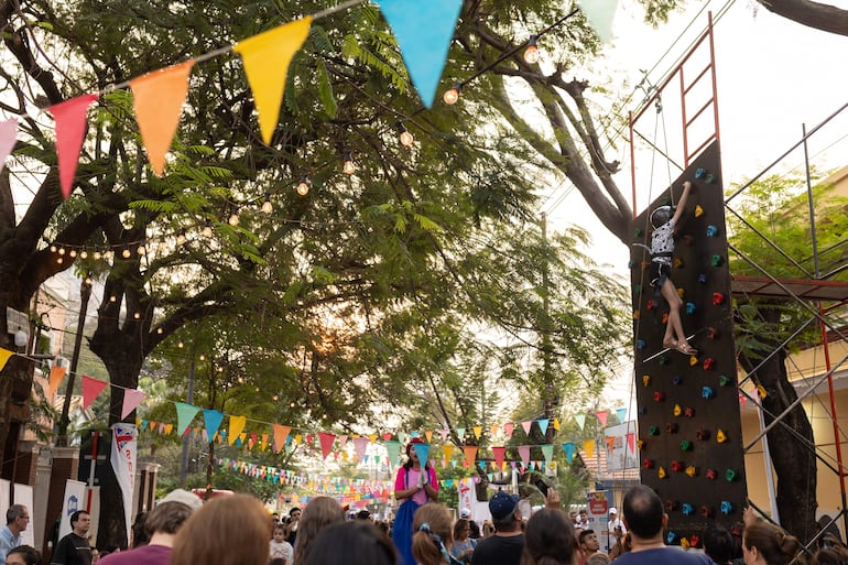 Mujer con blusa colorida anima al público, mientras un niño escala una pared en un ambiente festivo con banderines.