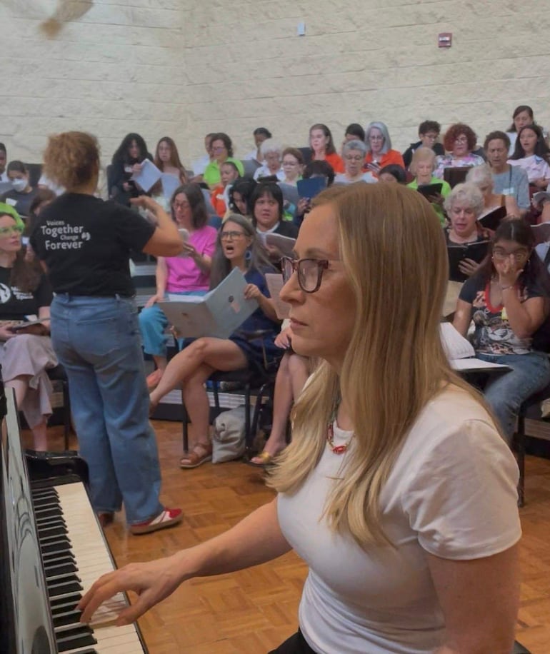 Mujer rubia con gafas toca el piano mientras guía a un grupo de personas vestidas con colores vibrantes en un salón.