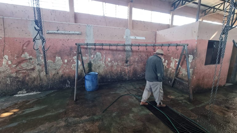 Hombre maduro con abrigo gris y pantalones claros camina por matadero con paredes desgastadas y ganchos colgantes.