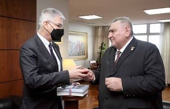 Marc Ostfield y Antonio Fretes se encuentran de pie en un despacho, ambos con trajes oscuros y expresiones serenas, intercambiando un objeto.