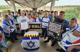 Hombre mayor con camiseta blanca 'LIGHT' y otro hombre con camiseta azul con estrella de David, rodeados de velas y banderas.