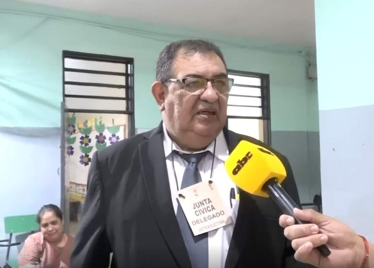 Inocencio Rodríguez, con traje oscuro y corbata, sostiene un micrófono amarillo mientras una mujer escucha en segundo plano.