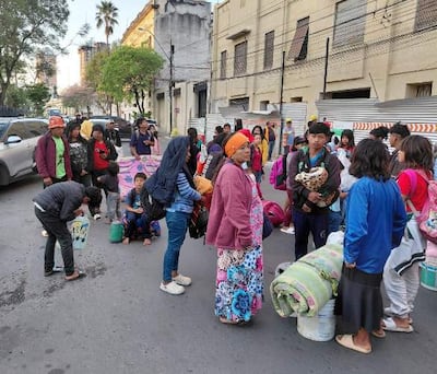 Veinte indígenas, en su mayoría adultos, se agrupan con ropas coloridas y mantas, esperando en la calle frente a edificios del Congreso.