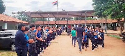 Grupo de niños con camisetas azules y educadores aplaudiendo en un camino central, con bandera paraguaya ondeando al fondo.