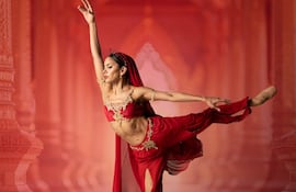 Bailarina en postura de danza clásica, vestida de rojo con dorados, en un fondo arquitectónico estilo palacio oriental.