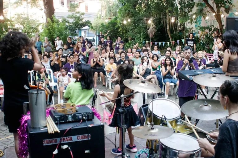 Dos niñas en el escenario: una cantando frente al micrófono y otra tocando la batería, con público aplaudiendo.