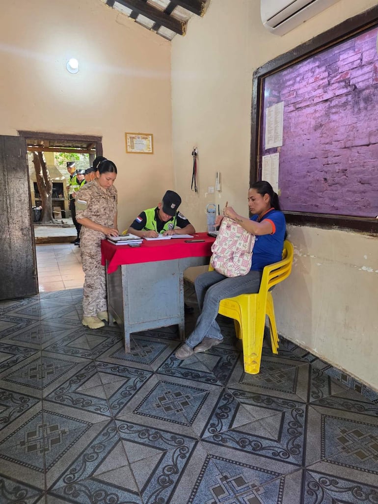 Mujer en uniforme camuflado de pie junto a hombre en uniforme negro y verde, otra mujer esperando sentada en una silla amarilla.