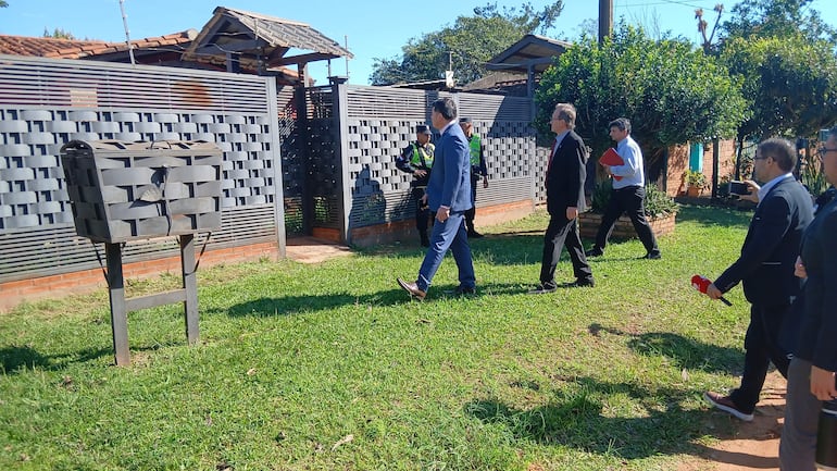 Seis personas, dos en trajes formales y una de seguridad, caminan por césped verde hacia la entrada de una propiedad con un muro moderno.