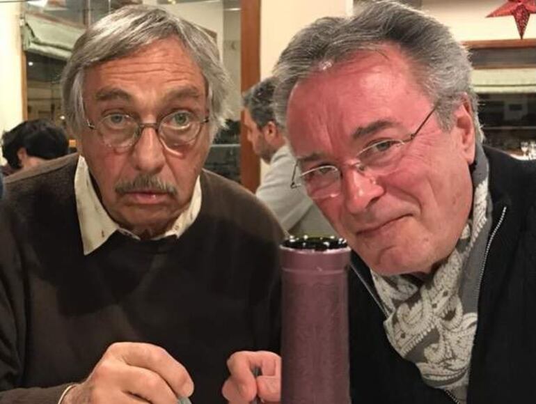 Dos hombres canosos sentados en una mesa, conversando seriamente. Uno lleva gafas, camisa oscura y el otro abrigo negro con bufanda clara.