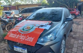 Automóvil gris estacionado en área policial con documentos sobre el techo, incluyendo 'EVIDENCIA' y letrero de 'Dirección de Policía Central'.