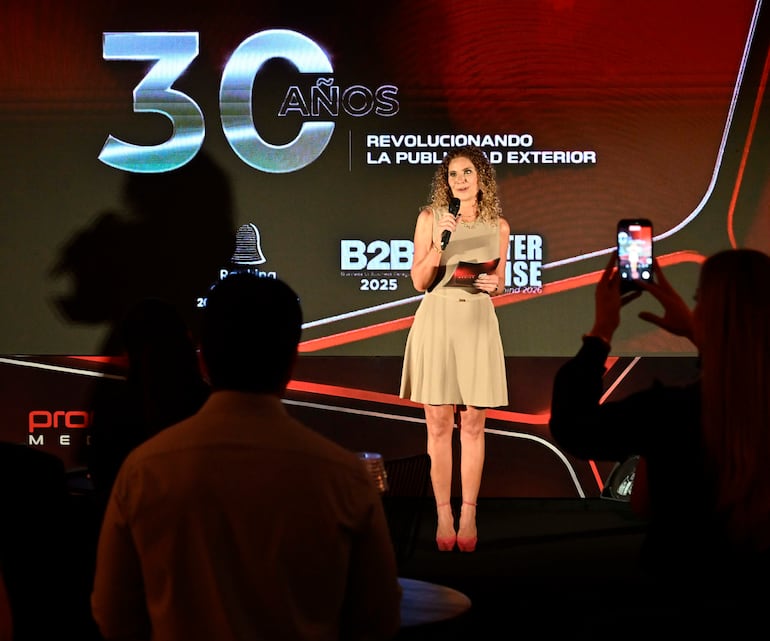 Mujer de pie en escenario con vestido claro, micrófono en mano, fondo con '30 AÑOS' y público atento.