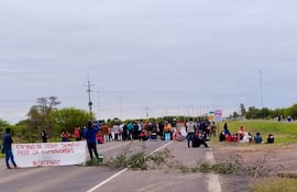 Grupo diverso de personas sosteniendo pancarta con mensaje claro, algunos sentados en la carretera, rodeados de vegetación.