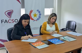 Dos mujeres en una mesa firmando documentos; una lleva suéter negro, la otra suéter amarillo, en ambiente institucional.