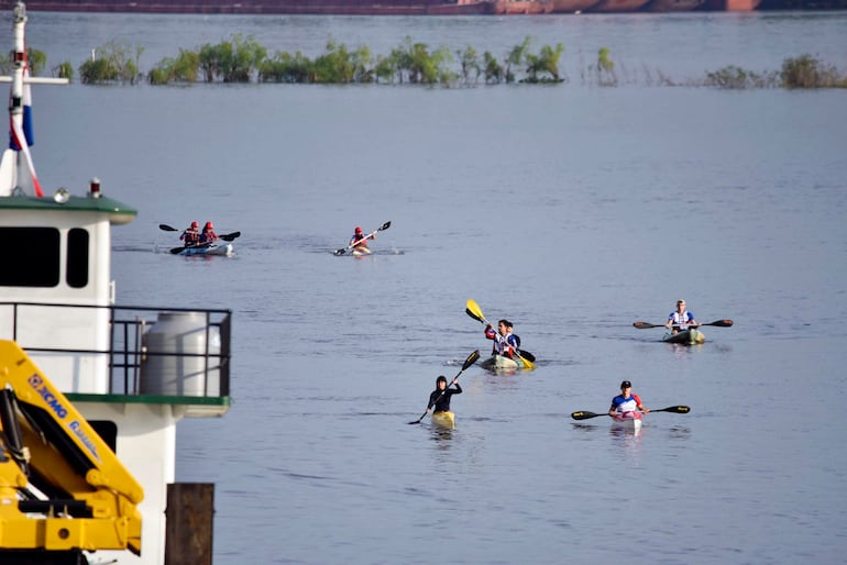 Seis remadores en kayaks de colores variados, algunos con chalecos rojos, navegan en un río rodeado de vegetación.